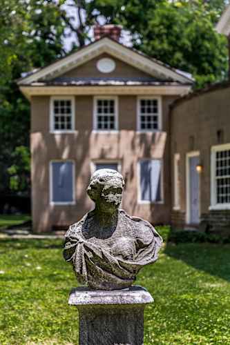A stone bust of a woman is in the foreground of a grassy area in front of a tan house with a black roof.