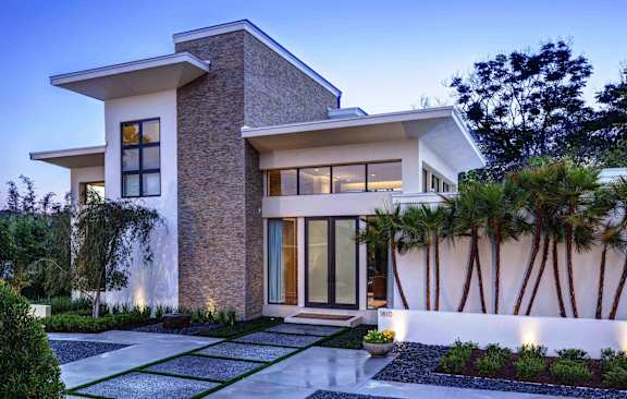 A modern house with a stone facade and a white wall with palm trees in front.