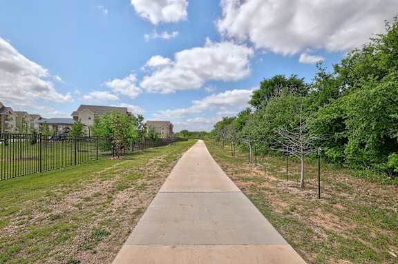 A long concrete pathway leads through a grassy area with trees and houses in the background.