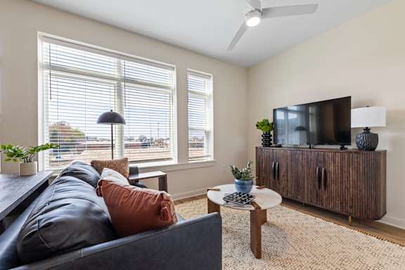 A living room with a grey couch, a wooden coffee table, a television, and a window with blinds at Hawkins Press Apartments, Charlotte, North Carolina