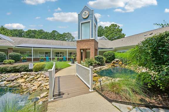 A small building with a clock tower is surrounded by a garden and a bridge.