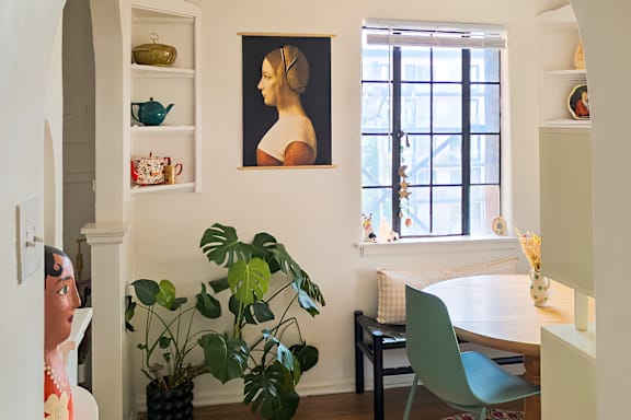 A woman is standing in a room with a potted plant, a painting, and a window.