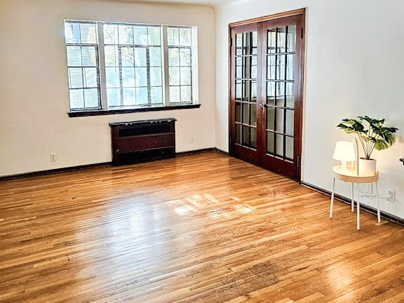 A room with a wooden floor, a piano, a table with a lamp and a plant, and a large window.
