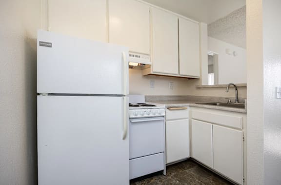 A white refrigerator stands in a kitchen with white cabinets and a white stove.