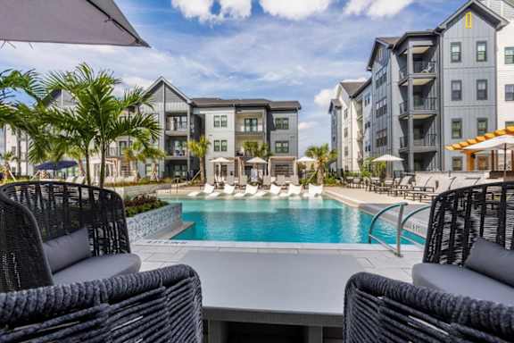 A pool surrounded by chairs and umbrellas with apartment buildings in the background.