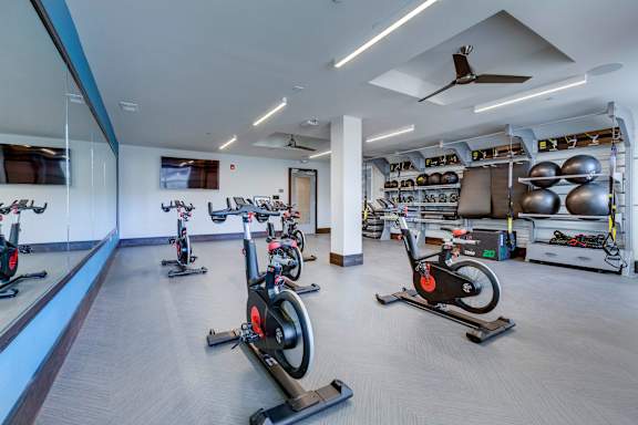 A gym with a row of stationary bikes and a fan on the ceiling.