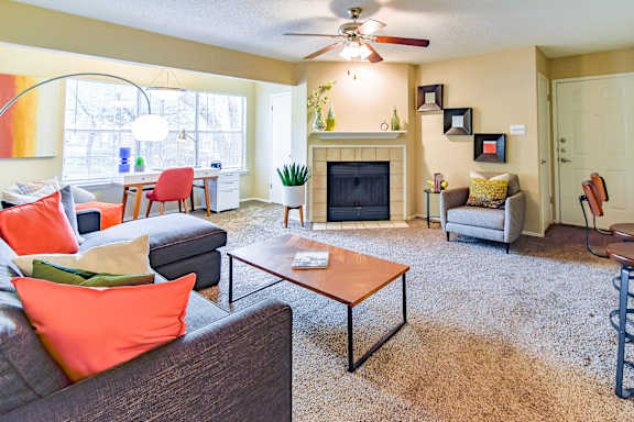 A living room with a grey couch, orange pillows, a coffee table, a fireplace, and a ceiling fan.