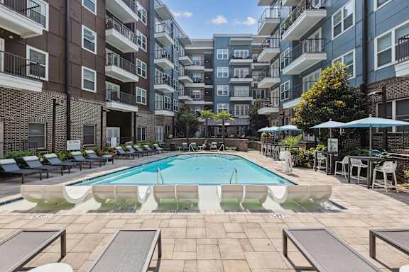 A swimming pool surrounded by lounge chairs and umbrellas in front of apartment buildings.