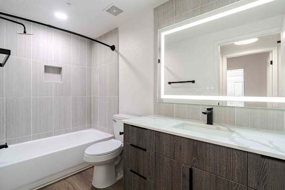 A white toilet sits in a bathroom with a white tub and a large mirror.