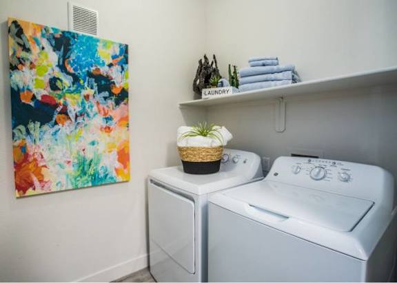 A laundry room with a washer and dryer, a basket of plants, and a colorful painting on the wall.