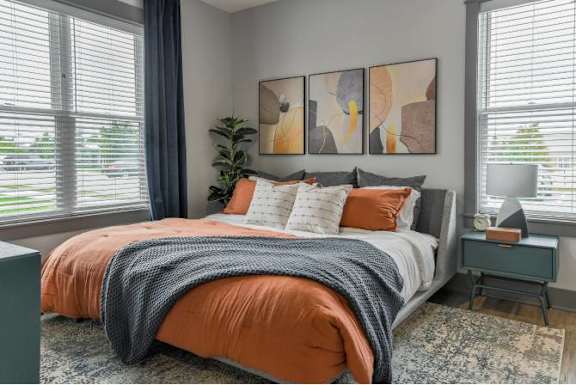 A bedroom with a bed covered in an orange comforter and a grey blanket, a small table with a lamp, and a plant on the windowsill.