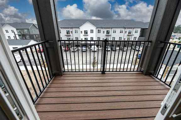 A balcony with a black railing and a wooden floor overlooks a parking lot and buildings.