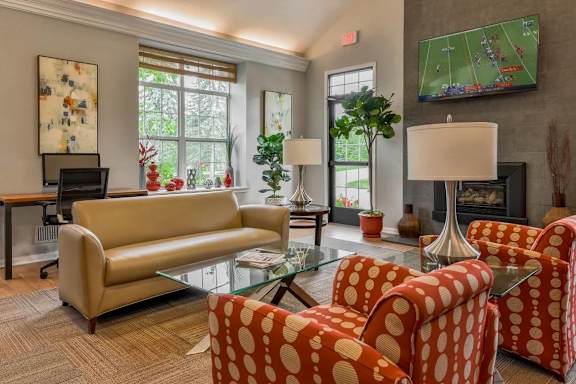 A living room with a tan couch, two red and white patterned chairs, a glass coffee table, and a television playing a football game.