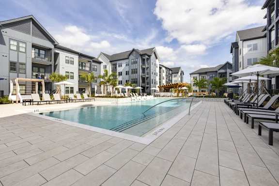 A large swimming pool surrounded by lounge chairs and umbrellas in front of apartment buildings.