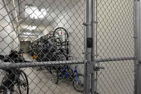A chain link fence with a gate is in front of a wall with a bunch of bicycles hanging on it.