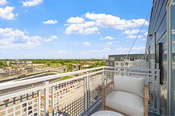 A balcony with a white chair and a table overlooking a parking lot and buildings.