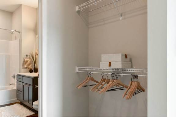 A white closet with clothes hanging and a white shelf with boxes on it.