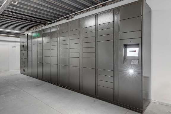 A row of tall, dark grey lockers are lined up in a room with a concrete floor and a white wall.
