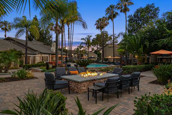 A patio with a fire pit surrounded by chairs and palm trees.
