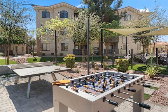 A foosball table is in the middle of a patio with a picnic table and a building in the background.