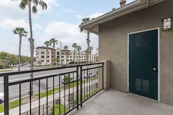 A balcony with a green door and a black railing overlooks a street with palm trees.