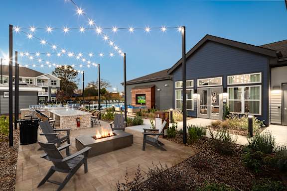 A patio area with a fire pit and chairs is surrounded by a building and string lights.
