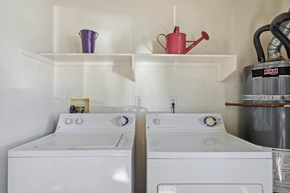 Two white washing machines are in a laundry room with a red watering can and purple bucket on the shelf above them.