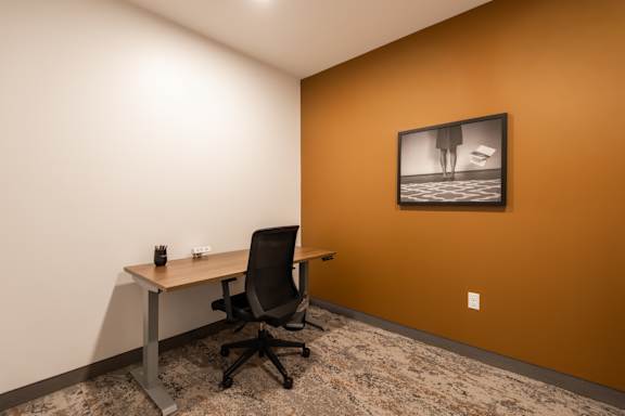 A black office chair is in front of a desk with a framed picture of a woman on the wall.
