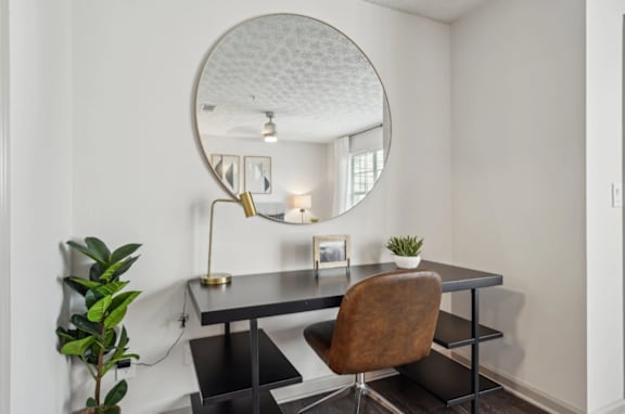 A round mirror hangs on a white wall above a black table with a brown chair and a green plant.