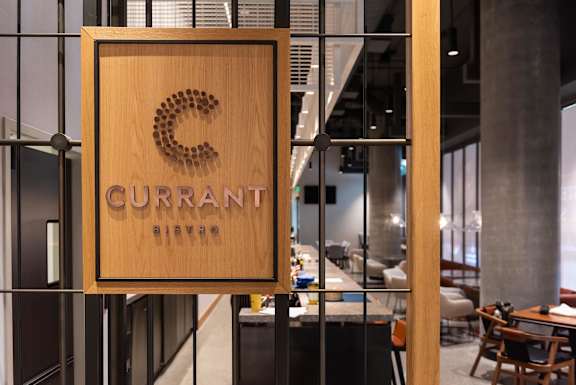 A wooden sign with the word "CURRANT" and a logo is hanging on a wall.
