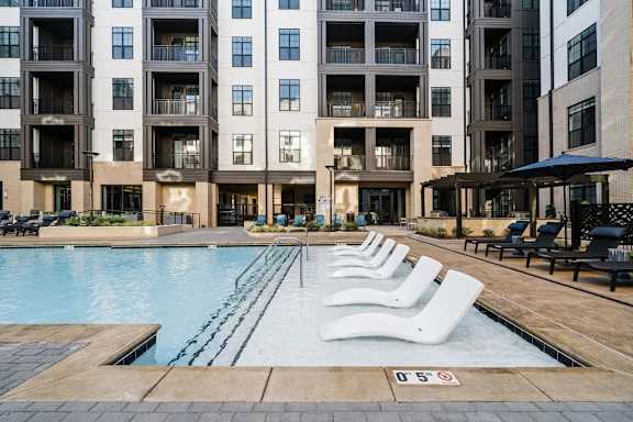 A swimming pool surrounded by lounge chairs in front of apartment buildings.