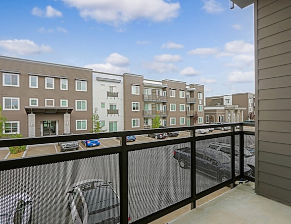 A balcony with a black railing overlooks a parking lot and apartment buildings.