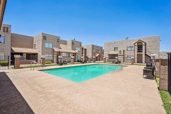 A swimming pool is surrounded by a concrete patio and a fence in front of a building.