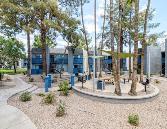 A modern building with blue doors and windows is surrounded by trees and a gravel area.