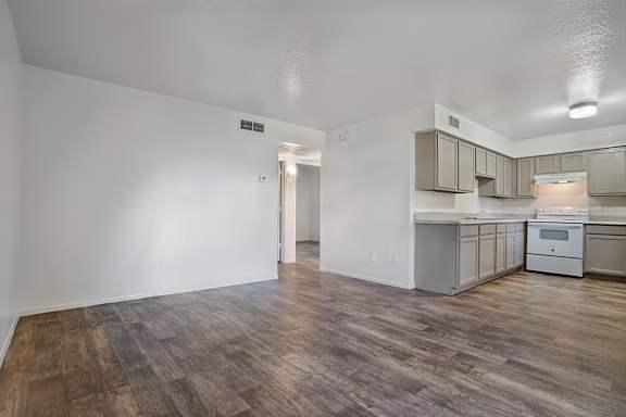 A room with wooden flooring and white walls, leading to a kitchen area.