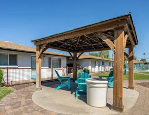A wooden pavilion with a white trash can and blue chairs in front of a house.