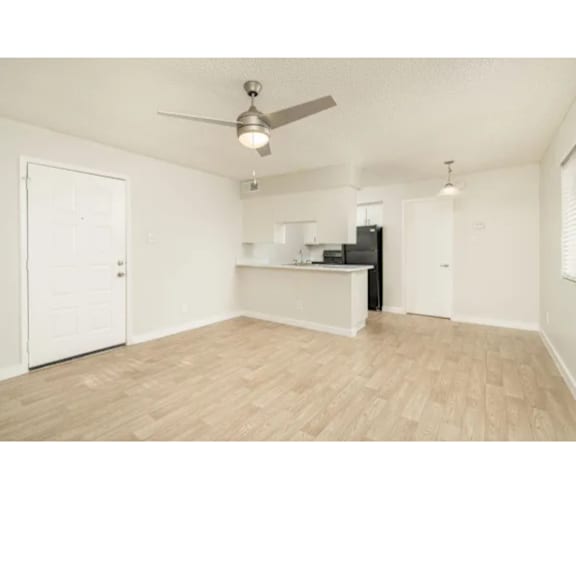 A room with a ceiling fan and light fixture, a small kitchenette, and a door on the left.