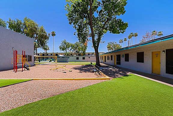 A playground with a slide, swings, and a tree in front of a building.