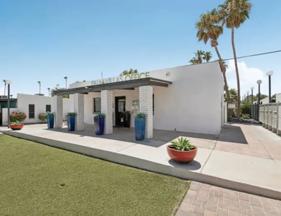 A white building with a black roof and a sign that reads "BATAVILAS OFFICE" is surrounded by a grassy area and a few potted plants.