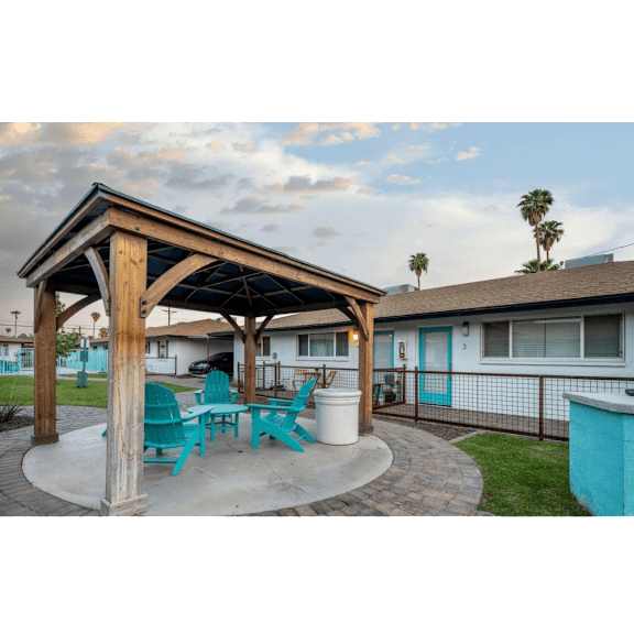 A wooden pergola with blue chairs and a white planter is in the foreground of a residential area.