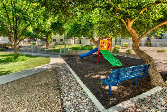 a playground with green and blue slides, a blue bench, and other play equipment under a tree-shaded area at Connect on Northern in Glendale, Arziona