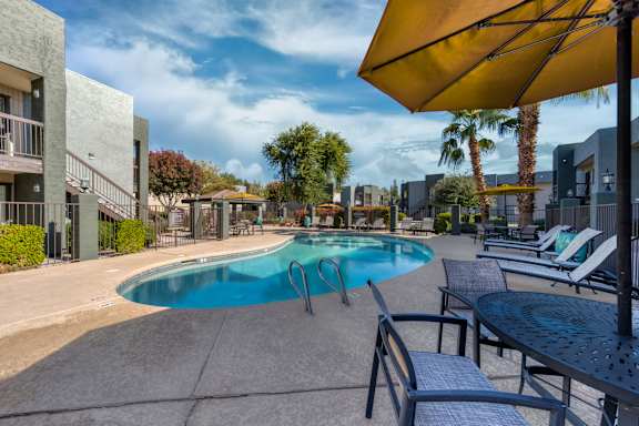 A pool area with a blue pool, sun loungers and a yellow umbrella.