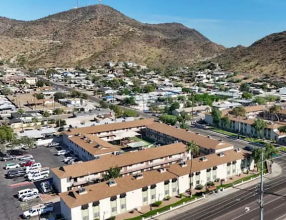 A large white building with a brown roof is surrounded by a parking lot and other buildings in a desert-like area.