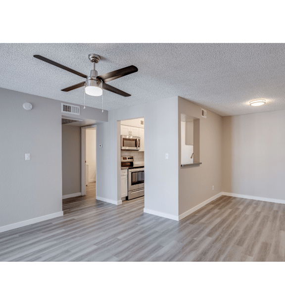 A room with a ceiling fan and a kitchen in the background.