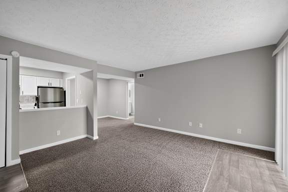 A spacious living room with grey carpeting and a kitchen area in the background.