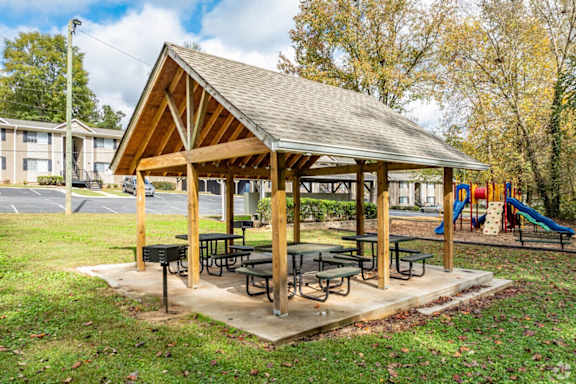 A wooden pavilion with a picnic table is surrounded by a playground and apartment buildings.