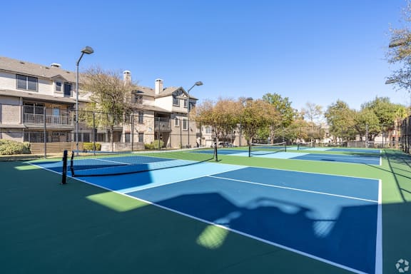 A tennis court with a blue surface and white lines is surrounded by apartment buildings.
