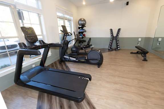 A treadmill, a stationary bike, and a rowing machine are in a room with wooden floors and windows.