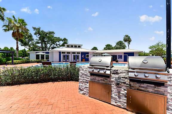 A house with a blue and white exterior is surrounded by greenery and has a brick patio with a grill.