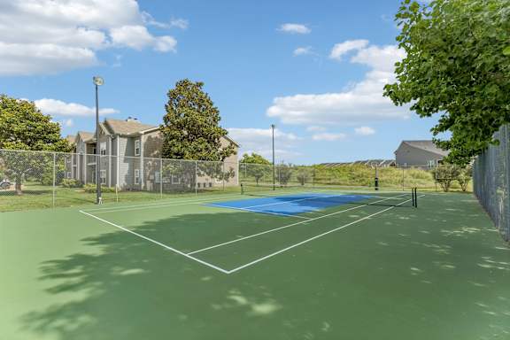 A tennis court with a blue surface and white lines, surrounded by a fence and houses in the background.